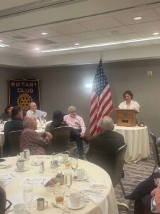 female standing behind a lectern with american flag nearbuy and people sitting around a table